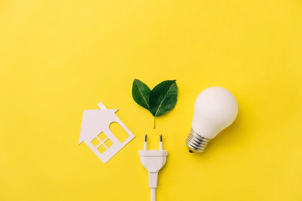 Concept for saving energy, eco-friendly, global warming. Creative top view flat lay of LED light bulb, electrical plug, leaves, paper house composition with copy space on yellow background