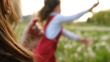 Gün batımında, neşeli anaokulu kızının arka planında parkta karahindiba üfleyen genç annenin yan görüntüsü. Yakın aile yaz tatilinde doğanın tadını çıkarır. Seçici odaklanma. Yavaş çekim