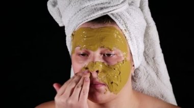 Evde kafasında beyaz havlu olan bir kadın kilden maske takıyor..