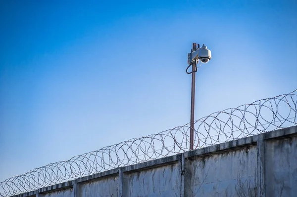 Close up view of security camera hanging among barbwire in prison or other guarded object with blue sky background. Modern ways of supervision. Using new technology in security and safety.