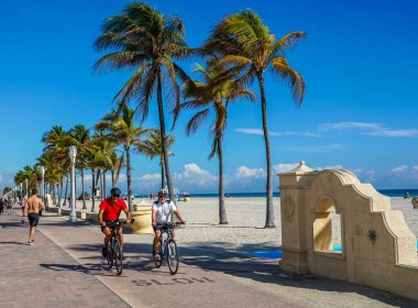 HOLLYWOOD BEACH, FLORIDA - JANUARY 29, 2020: Bicycle rider at the Hollywood Beach Broadwalk in South Florida. The promenade along the beach lined with palm trees and resorts is a popular tourist destination in Broward County clipart