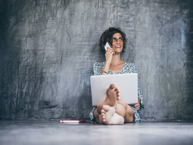 Kadın dizüstü bilgisayarla yerde oturuyor ve akıllı telefondan konuşuyor. 