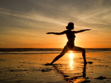 Gün batımında kumsalda yoga yapan genç bir kadın. 