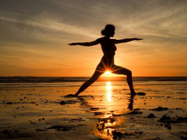 Gün batımında kumsalda yoga yapan genç bir kadın. 