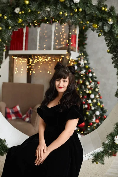 Плюс розмір моделі дівчини в інтер'єрі прикрашений на Новий рік. Модна пухка брюнетка в чорній вечірній сукні, з червоними губами і стильною зачіскою, позує з ялинкою на задньому плані . — стокове фото