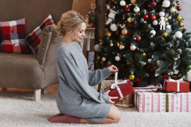 Örgü elbiseli, platin sarısı saçlı güzel bir kız Noel hediyesiyle birlikte iç kısımda oturuyor. Yeni yıl sabahında Noel ağacının yanındaki hediye paketini açan güzel bir sarışın.