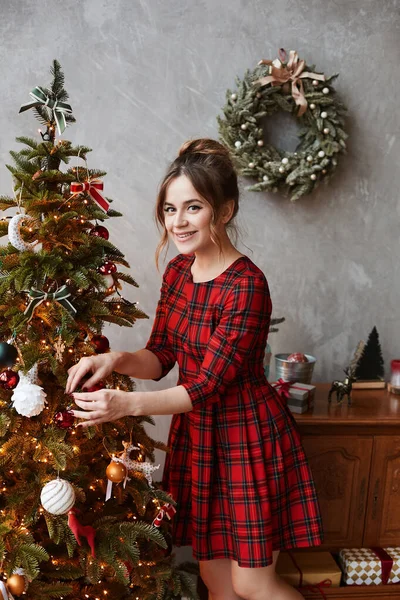 Gelukkig glimlachend model meisje met trendy kapsel in ruitvormige rode jurk versieren kerstboom. Jonge mooie vrouw in modieuze outfit poseren in de buurt van kerstboom en voor te bereiden op nieuwjaar te vieren — Stockfoto