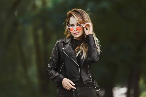 Fashionable beautiful and sensual blonde model girl in black leather jacket and jeans adjusting her stylish sunglasses and posing outdoors at the city street — Stock Photo, Image