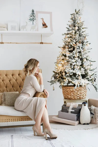 Gyönyörű platina szőke modell lány tökéletes test kötött ruhában pózol közel karácsonyfa belső. Fiatal karcsú modell nő divatos ruhában ül egy kanapén újévi belsőépítészet. — Stock Fotó