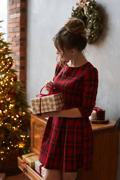 Модная девушка-модель с триумфальной прической в красном платье держит подарочную коробку возле елки. Молодая красивая женщина распаковывает рождественский подарок в интерьер, украшенный к Новому году. — стоковое фото
