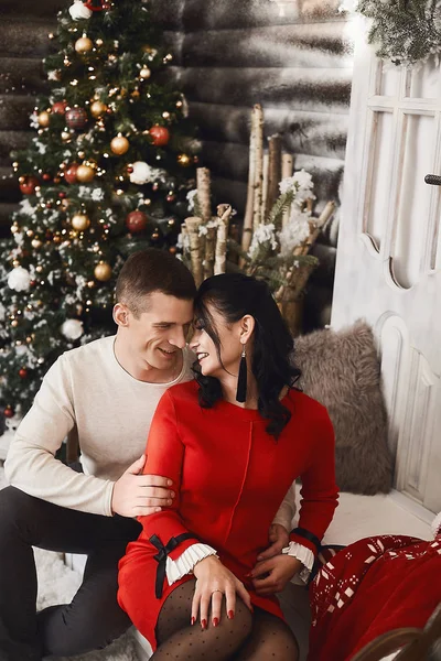 Jeune beau couple d'amoureux, heureuse femme brune en robe rouge et beau homme souriant posant à l'intérieur avec des lumières de Noël festives sur le fond. Bonne paire posant près de l'arbre de Noël . — Photo