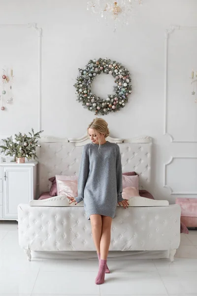 Fashionable model girl with platinum blond hair in knitted dress posing in bedroom decorated for Christmas. Young leggy woman with perfect slim body in cozy outfit posing in New year interior