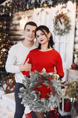 Birbirlerine aşık genç bir çift güzel dekore edilmiş bir Noel ağacının yanında poz veriyorlar. Güzel genç bir kadın ve elinde Noel çelengi tutan yakışıklı bir adam ve yeni yıl için iç dekorasyonda poz veriyor..