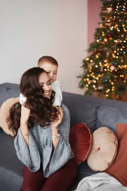 Genç, gülümseyen bir anne, küçük mutlu oğluna sarılıyor ve Noel arifesinde evde poz veriyor. Bulanık Noel ışıklarına karşı bebekli mutlu bir annenin portresi. Kış tatili kavramı