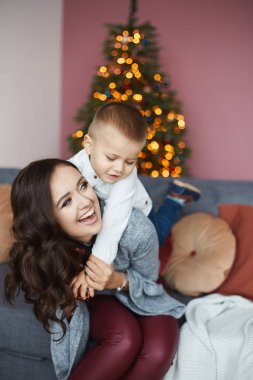 Genç, gülümseyen bir anne, küçük mutlu oğluna sarılıyor ve Noel arifesinde evde poz veriyor. Bulanık Noel ışıklarına karşı bebekli mutlu bir annenin portresi. Kış tatili kavramı