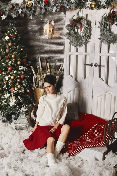 Wspaniała młoda kobieta w czerwonej eleganckiej spódnicy i białym swetrze siedzi na schodach w pobliżu choinki w przytulnym wnętrzu. Modny model dziewczyna w przytulnym stroju we wnętrzu z dekoracji noworocznych — Zdjęcie stockowe