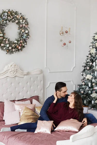 Sweet couple hugging each other on the bed in bedroom decorated for Christmas. Handsome man and pretty model girl in cozy winter clothes spending together winter and New year holidays