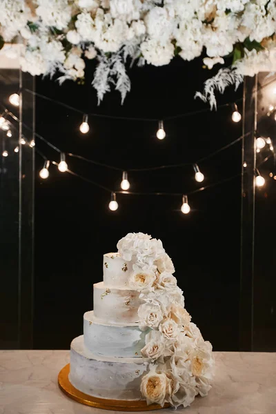 White wedding cake with peonies and roses flowers on the night ceremony outdoors. Extra large three-tiered wedding cake with white buttercream frosting and fresh white flowers. Luxury birthday cake