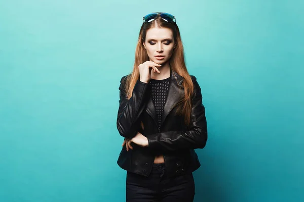 Stylish model girl with modish makeup in leather jacket and sunglasses isolated at light blue background. Fashionable young woman with bright makeup, dressed in rock style, posing over blue background — Stock Photo, Image
