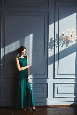 Yeşil gece elbisesi içinde, modaya uygun saç stili olan güzel genç bir kadın mavi duvarda, güneş ışığı ve gölgelerle dikiliyor. Model elbiseli kız, eski moda iç mekanlarda güneşli bir günün tadını çıkarıyor.