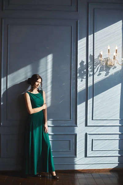 Beautiful young woman with modish hairstyle in green evening dress stands with closed eyes at blue wall with sunlight and shadows. Model girl in a modish outfit enjoys a sunny day in vintage interior — 스톡 사진