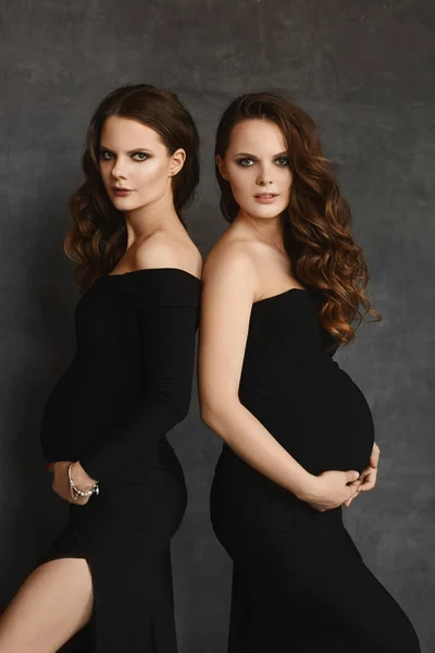 Toekomstige moeders houden handen op de buik en kijken in de camera. Twee jonge zwangere vrouwen in zwarte jurken met grote buiken. Mooie zwangere tweeling die dicht bij elkaar blijven op de donkere achtergrond. — Stockfoto