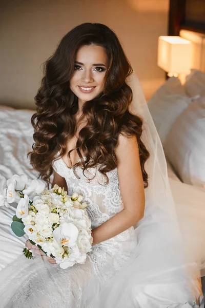 Mulher morena jovem com penteado de casamento em um vestido de renda mantém um buquê de flores de falaenopsis, rosas e peônias nas mãos. Menina modelo impressionante em vestido de noiva segurando buquê de flores exóticas — Fotografia de Stock