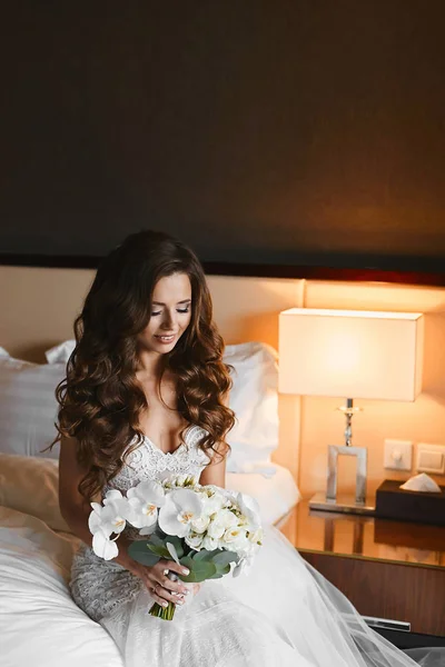 Mulher morena jovem com penteado de casamento em um vestido de renda mantém um buquê de flores de falaenopsis, rosas e peônias nas mãos. Menina modelo impressionante em vestido de noiva segurando buquê de flores exóticas — Fotografia de Stock