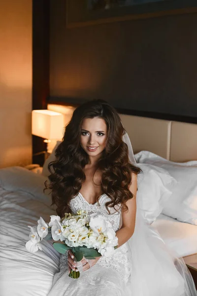 Mulher morena jovem com penteado de casamento em um vestido de renda mantém um buquê de flores de falaenopsis, rosas e peônias nas mãos. Menina modelo impressionante em vestido de noiva segurando buquê de flores exóticas — Fotografia de Stock