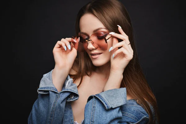 Ung modell kvinna i coola rosa solglasögon och denim jacka poserar på svart isolerad bakgrund. Attraktiv tjej med ljus makeup i jeans jacka ler och poserar i studio. Tillbehör och makeup — Stockfoto