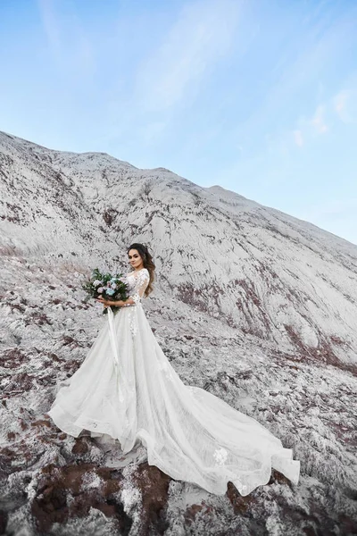 Ein wunderschönes Model-Mädchen, junge Braut, in einem modernen Brautkleid, das Brautstrauß hält und auf einem weißen Felsen mit blauem Himmel im Hintergrund posiert. Hochzeitsmode-Konzept. Kopierraum an der Spitze — Stockfoto