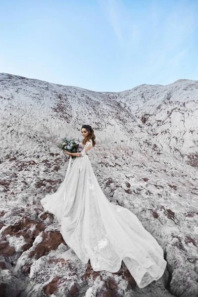 Eine schöne junge Frau in einem modernen Brautkleid hält Brautstrauß und posiert an einem sonnigen Frühlingstag auf einem Berg mit blauem Himmel im Hintergrund. Hochzeitsmode-Konzept. Kopierraum an der Spitze — Stockfoto