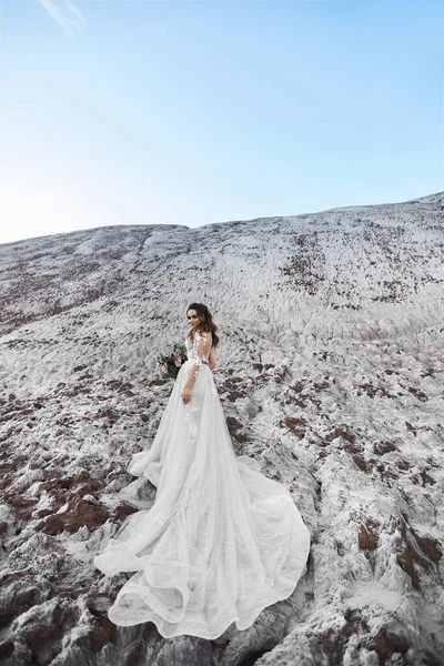 Una bella giovane donna in un abito da sposa moderno mantenendo bouquet da sposa e in posa su una montagna con cielo blu sullo sfondo in una giornata estiva soleggiata. Concetto di moda matrimonio. Copia lo spazio in alto — Foto Stock