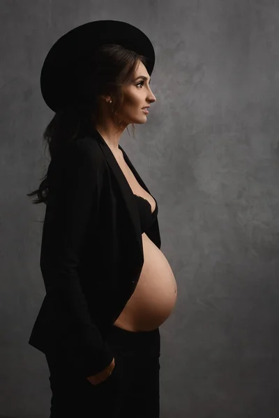 High fashioned jonge zwangere vrouw met een trendy hoed en een modieuze zwarte outfit met blote buik staat zijwaarts en kijkt weg van de grijze achtergrond. Concept van zwangerschap mode — Stockfoto