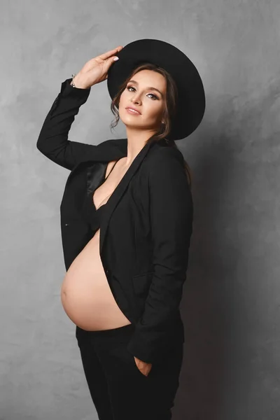 Mulher grávida feliz bonita com barriga nua, em jaqueta desabotoada e roupa interior preta, ajustando seu chapéu modish e sorrindo para a câmera. Futura mãe, um retrato de uma jovem grávida em lingerie . — Fotografia de Stock