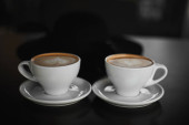 Dva bílé šálky kávy Cappuccino s mléčnou pěnou ve tvaru srdce. Boční pohled na dva šálky latté kávy se srdeční postavou na mléčné pěně na černém pozadí. Životní styl