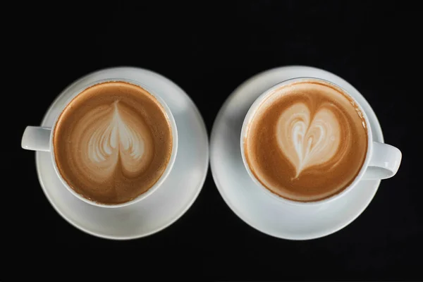 Duas xícaras brancas de café Cappuccino com espuma de leite em forma de coração. Vista superior em duas xícaras de café latte com figura do coração em espuma de leite isolado em fundo preto. Conceito de pequeno-almoço. Vista de cima — Fotografia de Stock