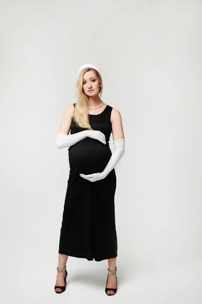 Beautiful pregnant woman in black evening dress and long white gloves touching her belly and posing on a white background, isolated. High fashioned portrait of young mother anticipation of the baby.