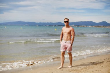 Handsome shirtless athletic young guy walking on the beach by the waterline. Young muscular man with perfect body in shorts and sunglasses walking by the sea coast. Travel fashion. Sport lifestyle