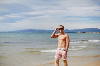 Handsome shirtless athletic young guy walking on the beach by the waterline. Young muscular man with perfect body in shorts and sunglasses walking by the sea coast. Travel fashion. Sport lifestyle