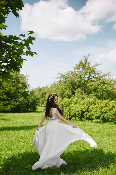 Beyaz uzun elbiseli, mutlu genç bir kadın bahar bahçesinde eğleniyor. Çiçek desenli düğün çelengi takmış genç gelin yaz güneşli bir günde etrafta dönüyor ve doğada poz veriyor. — Stok fotoğraf