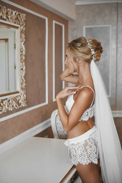 Giovane donna con corpo sottile e capelli biondo platino in posa vicino allo specchio in interni d'annata. Ragazza modello bionda con corpo sottile e acconciatura da sposa in lingerie di pizzo in posa all'interno . — Foto Stock