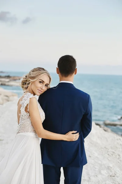 Красивая молодая блондинка в белом кружевном платье опирается на красивого мужчину в стильном синем костюме на белом камне у побережья Адриатического моря. Модель девушки в свадебном платье со стильным женихом — стоковое фото