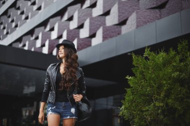 Deri ceketli, dalgalı saçlı, siyah şapkalı ve kot şortlu modern binanın yanındaki sokakta duran model bir kız. Moda kıyafetleri. Her gün bak. Sokak modası.