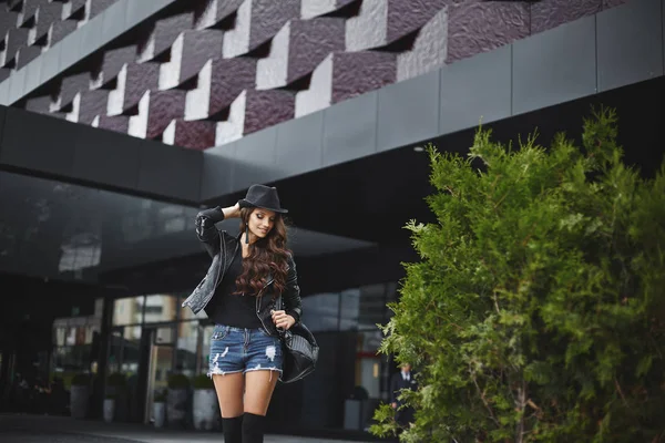 Young woman in denim shorts, black hat and leather jacket with black leather backpack walking in the street of European city. Trendy casual outfit. Everyday look. Street fashion. — Stock Photo, Image