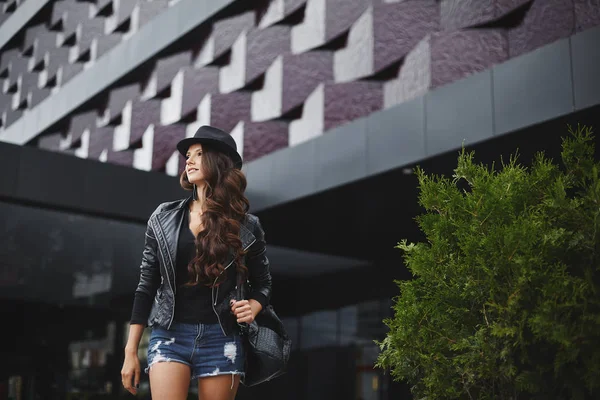 Modish model girl with wavy hair in a leather jacket, black hat and jeans shorts with a stylish backpack stands on the street near modern building. Trendy casual outfit. Everyday look. Street fashion.