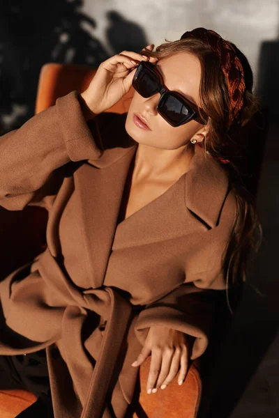 Модное фото красивой молодой женщины в модных солнцезащитных очках и пальто в кресле в интерьере. Модель девушки в весеннем наряде позирует в интерьере. Городской образ жизни Женская мода. Портрет крупного плана — стоковое фото