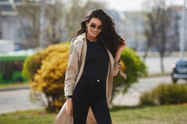 Bej rengi yağmurluk ve güneşli bahar gününde şehir sokaklarında poz veren siyah kotlu mükemmel vücutlu baştan çıkarıcı genç bir kadın. Şık bahar kıyafetleriyle esmer model kız şehir arka planında poz veriyor.