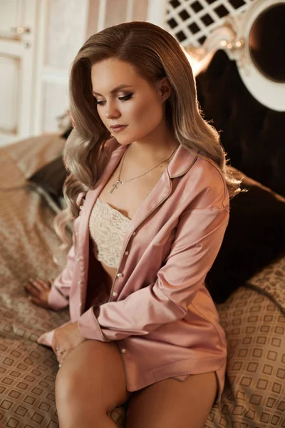 Retrato de mulher jovem com cabelo loiro de platina em pijama rosa de cetim na cama. Menina modelo com rosto de pele perfeita e penteado ondulado posando em um quarto de um apartamento de hotel de luxo — Fotografia de Stock