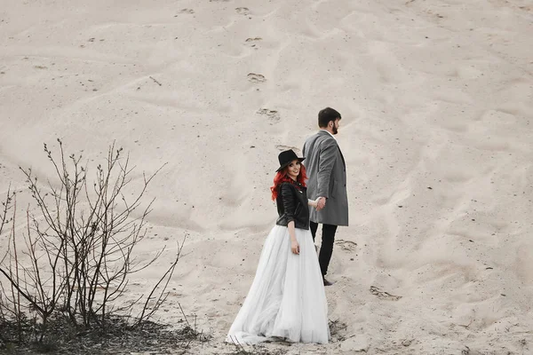 Joven novia en un vestido blanco, chaqueta de cuero negro y sombrero siguiendo al novio guapo joven y mirando hacia atrás a la cámara. Una feliz pareja de recién casados posando en el desierto. Concepto de amor y matrimonio — Foto de Stock